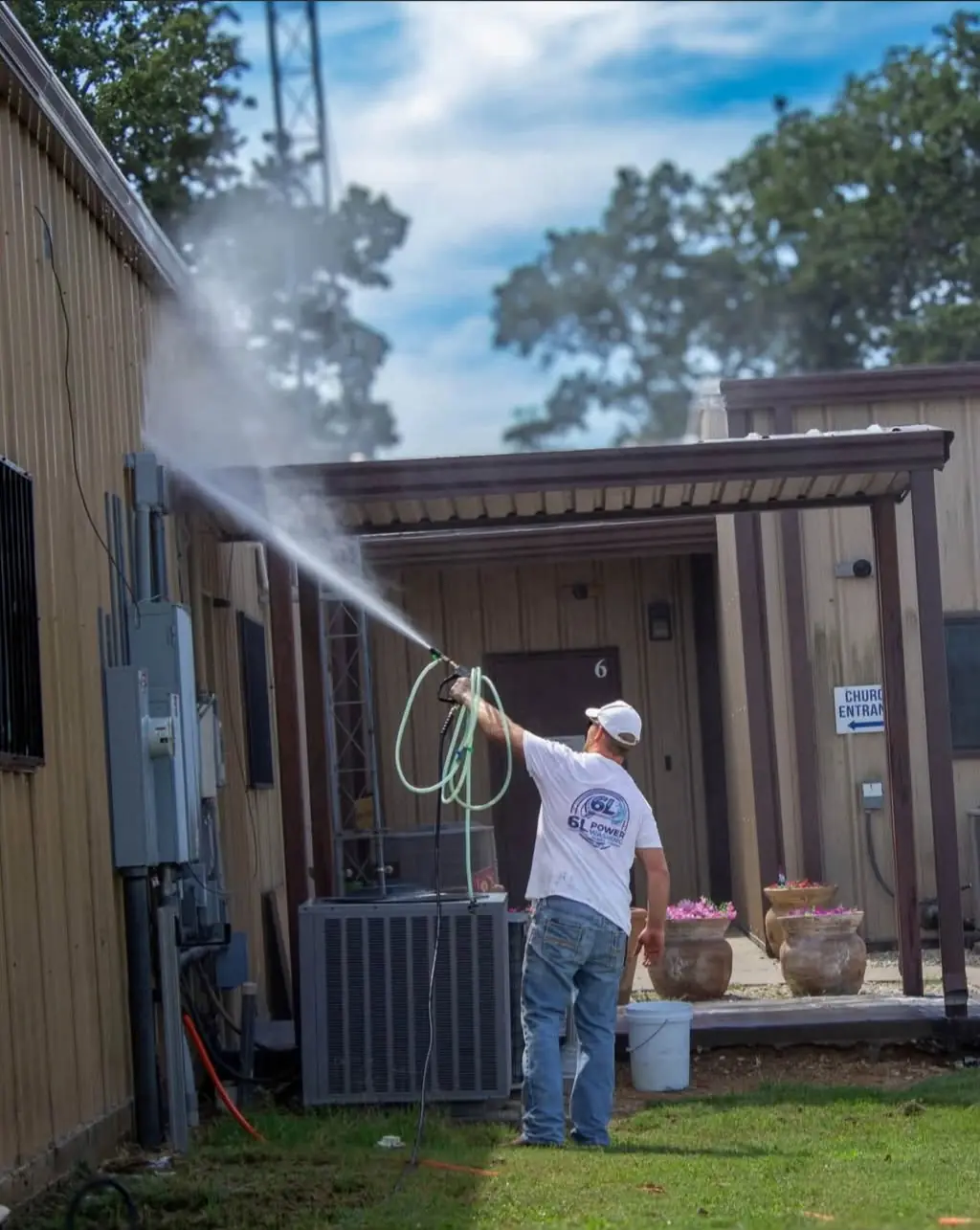 Man in a 6L Power Washing T-shirt spraying the upper wall of a wood-sided building with a pressure washer, a long arc of water against the sky